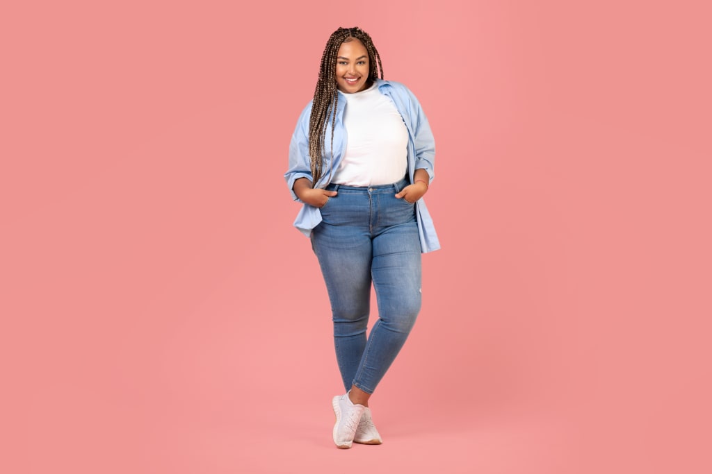 Woman with an Apple Body Shape stands against a pink background, wearing a light blue shirt, white t-shirt, blue jeans, and white sneakers, hands in her pockets and smiling.